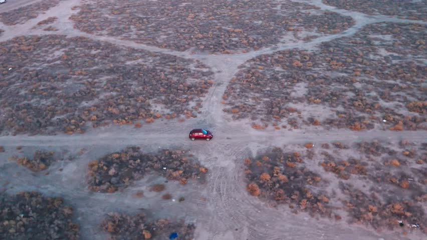 High aerial of a red car at a dusty desert crossroads amid scrub and branching trails at twilight—cinematic road-trip off-road establishing plate; empty landscape.