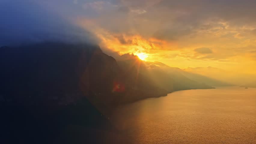 Golden sunset over Lago d’Iseo, with soft light spilling across mountain ridges and the calm lake below, captured from an elevated cinematic aerial perspective.