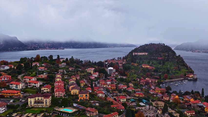 Descending drone shot over Bellagio on Lake Como, showing colorful hillside homes, the peninsula, calm water and snowy mist covering the surrounding mountains in a wintery, atmospheric scene.