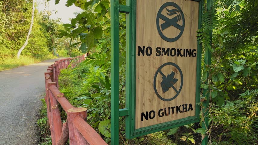 Static shot of a forest pathway signboard warning No Smoking and No Gutkha, mounted beside a quiet road with greenery and wooden railing nearby path.