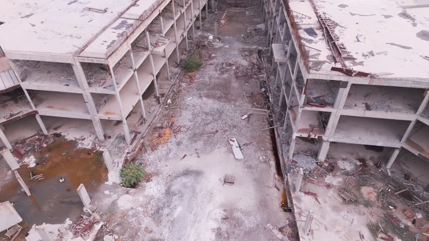 Drone flight down a deserted street in an unfinished building complex. Weeds grow through pavement cracks between concrete skeletons, creating a post-apocalyptic ghost town atmosphere.