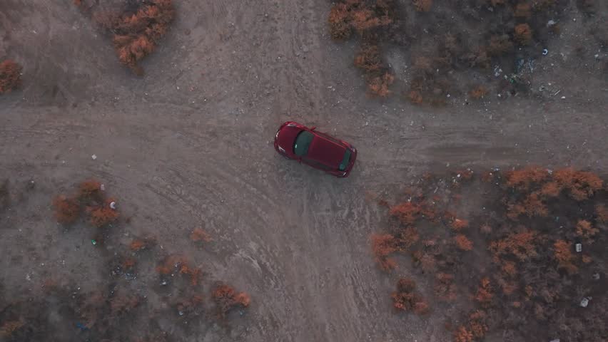 Overhead drone of a red car driving along a dusty desert track at twilight amid dry scrub and forked trails—cinematic travel and off-road establishing shot; empty landscape.