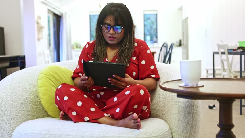 Indian woman in red pajamas with white polka dots sitting comfortably on a modern couch in her living room, actively using a tablet for entertainment, work or online shopping
