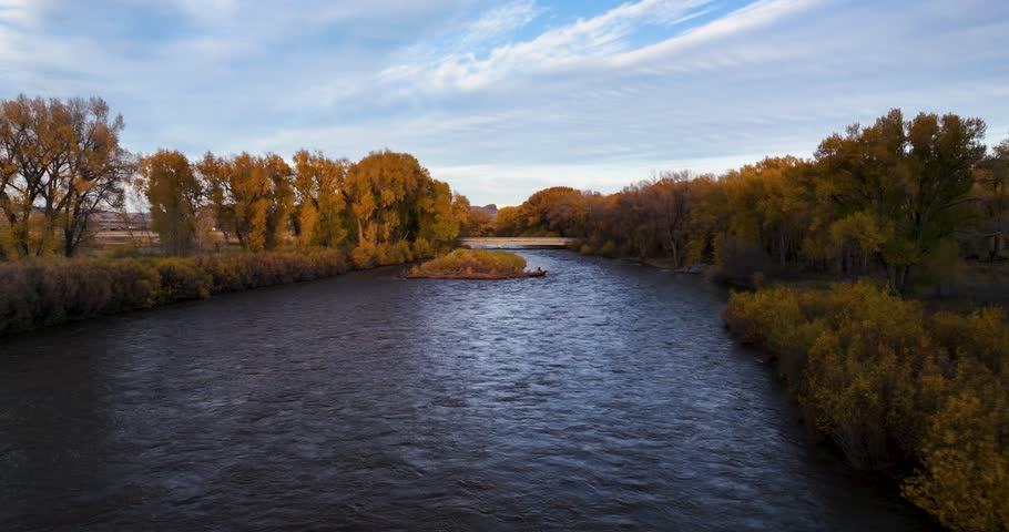 Aerial drone shot tracking low over a bridge crossing the Animas River, Colorado, with vibrant autumn foliage, bathed in warm sunset light, capturing the serene beauty of fall along the river.