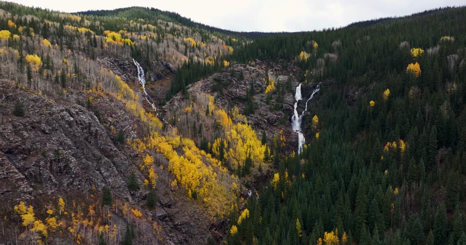 A 4K, forward tracking fly-over drone shot of a rushing mountain waterfall in Colorado during autumn, with lush green foliage on one side and vibrant yellow and orange fall colors on the other.