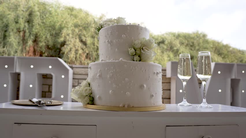 A lovely two-tier wedding cake adorned with white flowers stands elegantly on a table. Two champagne glasses sit nearby, creating a romantic atmosphere for the celebration.