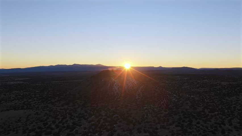 Wide drone shot of a solitary volcanic cinder cone rising from the high desert plains. The low-angle sun creates a dramatic silhouette, highlighting the rugged texture of the New Mexico landscape.