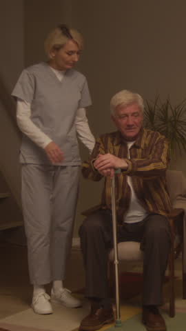 Vertical slowmo shot of middle-aged Caucasian female caregiver gently supporting senior Caucasian man using walking stick to move along in homey assisted living facility