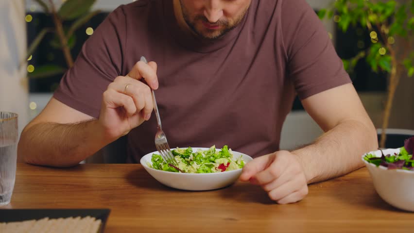 Unhappy thoughtful man picking at his plate sitting at table healthy vegetable salad bowl for diet dinner. Guy mixing salad with fork bad appetite, sad expression refuses to eat greens healthy eating.