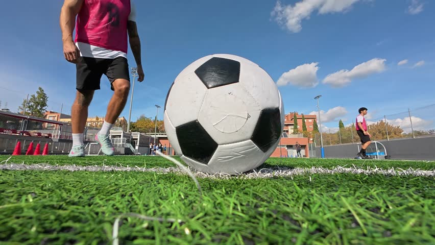 Low angle perspective of a football player kicking a ball on a turf field. The athlete performs a powerful shot during a training session