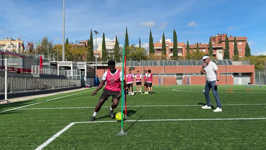 Young athletic man skillfully dribbling a soccer ball around a training pole on a green turf field. Professional coach observing his student