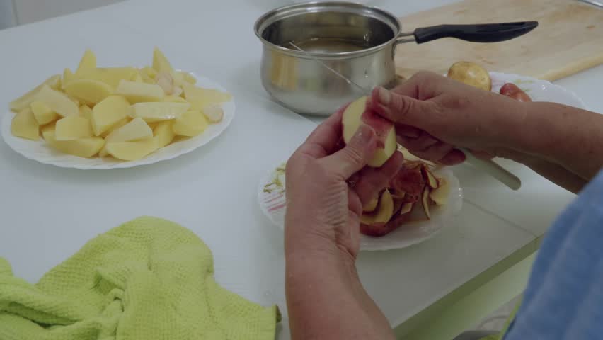 Close up of hands in blue shirt peeling red skin potato with small knife, removing peel strips over white floral plate, with sliced potatoes and saucepan nearby.