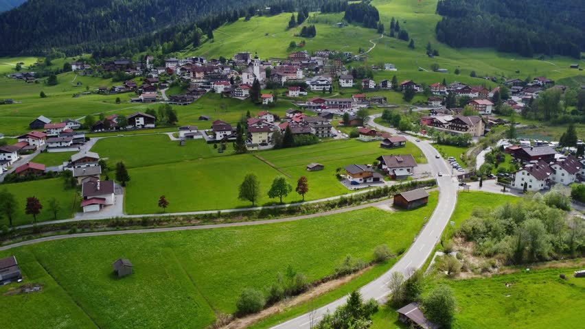 Aerial view of a scenic alpine village with a riverside campsite and traditional houses in Tyrol, Austria.The scene is surrounded by lush green meadows, pine forests, and winding roads, showcasing the idyllic rural lifestyle and tourism in the Tyrol region.