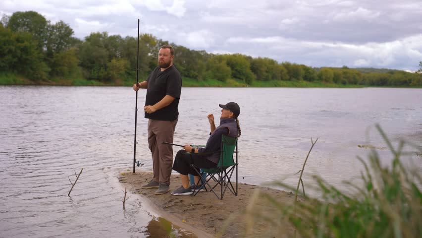 Young couple spending quality time together, sitting on folding chairs by the river and preparing their fishing rods for a day of angling, enjoying a quiet weekend getaway in nature