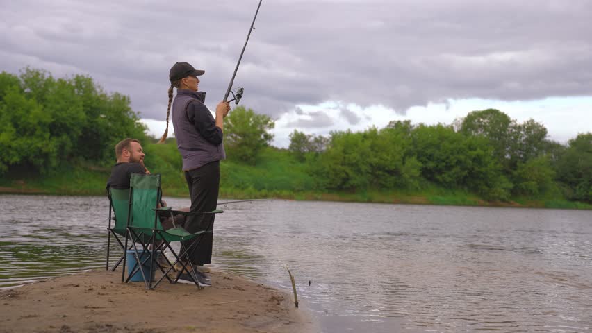 Young woman fishing with a spinning rod from the river bank and her boyfriend sitting nearby on a camping chair enjoying his outdoor recreation on a cloudy day