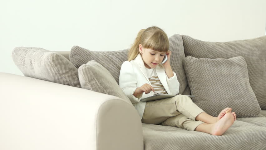 Little girl sitting on the couch with a tablet and talking on the phone
