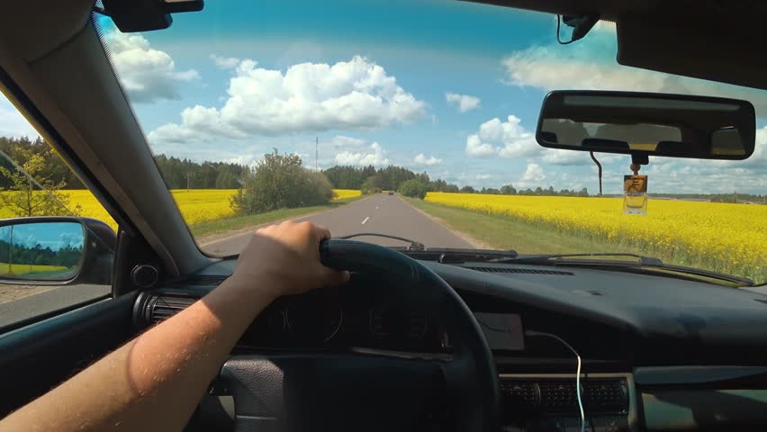 Pov of a person driving a car on a country road passing vibrant yellow rapeseed fields under a beautiful blue sky with white clouds, showing the concept of a scenic road trip in summer