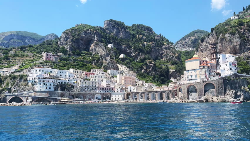 View from sailing boat on beautiful Atrani city along Amalfi coastline, picturesque old colourful buildings, tourists sea beach and blu water waves, charming panoramic view of Atrani city during the