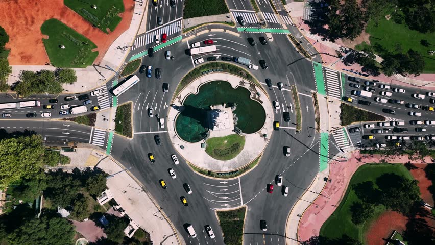 Aerial cinematic drone shot capturing the Monumento a los Españoles roundabout—officially known as the Monument to the Carta Magna and the Four Regions of Argentina—in the heart of Buenos Aires, Argentina. Filmed from a perfectly centered top-down perspective, the drone slowly ascends, revealing the symmetrical geometry of the traffic circle, the turquoise pools surrounding the monument, and the complex multi-lane flow of vehicles moving through one of the city’s busiest intersections. The shot highlights the contrast between the intense urban traffic and the lush green areas of Palermo that surround the avenue, including tree-lined paths, landscaped gardens, and vibrant red walking trails. The bright midday sunlight enhances the clarity, shadows, and architectural details, showcasing the monument’s sculpted figures and the characteristic urban layout of Buenos Aires. This footage is ideal for projects related to travel, tourism promotion, South American cities, transportation systems, traffic infrastructure, urban planning, aerial cinematography, and lifestyle content showcasing iconic landmarks in Argentina.