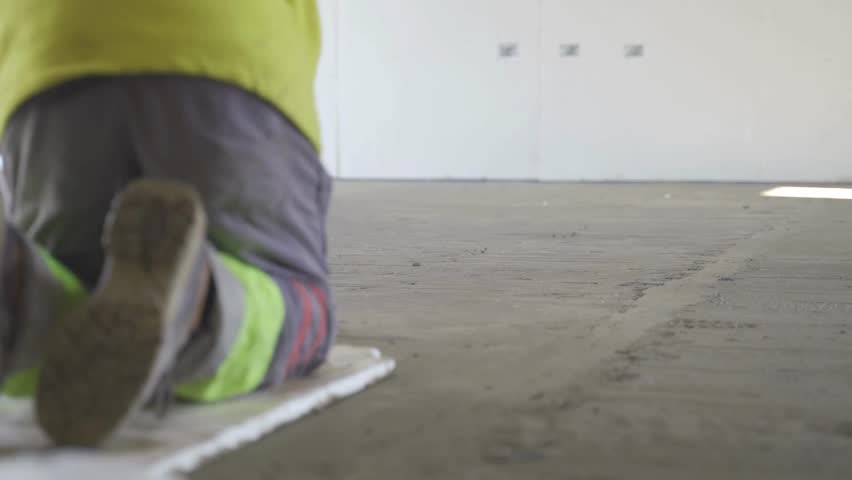A static view shows a laborer in a neon jacket and grey pants manually leveling a plaster floor with a flat trowel, highlighting precision, hard work, and hands-on craftsmanship.