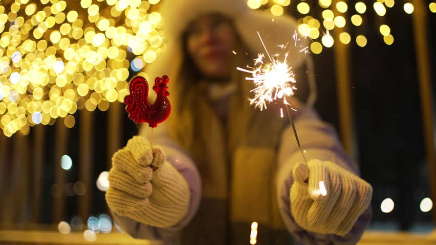 Festive woman in winter clothes holding a burning sparkler and a red cockerel lollipop at a christmas market, with bright golden bokeh lights creating a magical atmosphere in the background