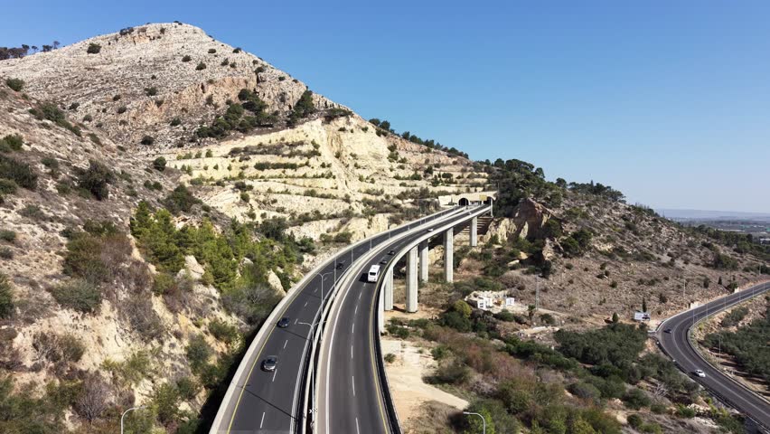 Aerial drone view of a highway bridge leading into twin road tunnels built through a rocky hillside in northern Israel. Cars drive toward the tunnel entrance surrounded by mountains, pine trees and open landscape, showing modern transportation infrastructure and civil engineering. This location is near Afula in the Jezreel Valley region and is part of the Highway 6 Menashe Tunnels area in Israel.