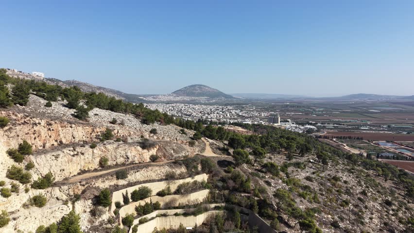 Aerial drone view of a highway bridge leading into twin road tunnels built through a rocky hillside in northern Israel. Cars drive toward the tunnel entrance surrounded by mountains, pine trees and open landscape, showing modern transportation infrastructure and civil engineering. This location is near Afula in the Jezreel Valley region and is part of the Highway 6 Menashe Tunnels area in Israel.