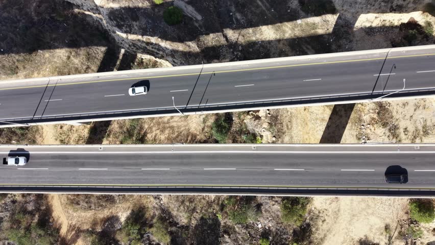 Aerial drone view of a highway bridge leading into twin road tunnels built through a rocky hillside in northern Israel. Cars drive toward the tunnel entrance surrounded by mountains, pine trees and open landscape, showing modern transportation infrastructure and civil engineering. This location is near Afula in the Jezreel Valley region and is part of the Highway 6 Menashe Tunnels area in Israel.