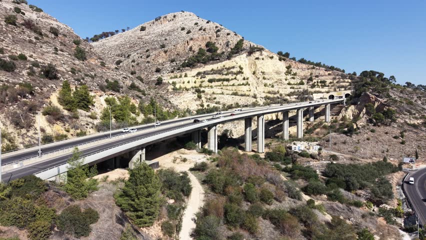 Aerial drone view of a highway bridge leading into twin road tunnels built through a rocky hillside in northern Israel. Cars drive toward the tunnel entrance surrounded by mountains, pine trees and open landscape, showing modern transportation infrastructure and civil engineering. This location is near Afula in the Jezreel Valley region and is part of the Highway 6 Menashe Tunnels area in Israel.