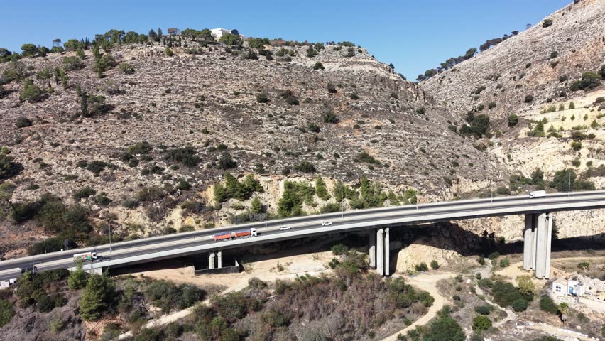 Aerial drone view of a highway bridge leading into twin road tunnels built through a rocky hillside in northern Israel. Cars drive toward the tunnel entrance surrounded by mountains, pine trees and open landscape, showing modern transportation infrastructure and civil engineering. This location is near Afula in the Jezreel Valley region and is part of the Highway 6 Menashe Tunnels area in Israel.