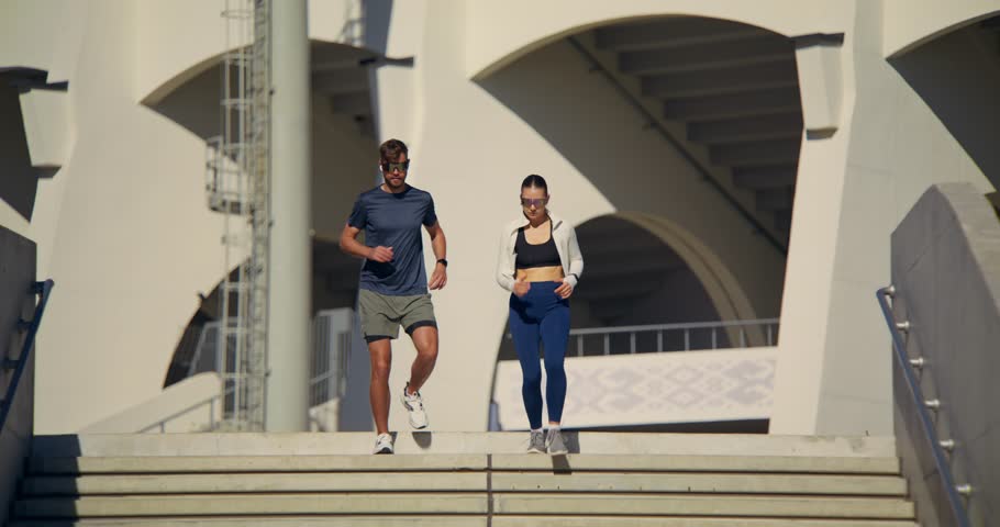 A sporty man and a woman are running in the city on a sunny day. They run down the city stairs