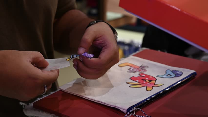 Bangkok, Thailand - 18th December 2025: Man manually printing alphabets on a pouch using an industrial printing machine, showcasing hands-on packaging, craftsmanship, manufacturing process, and small-scale production.