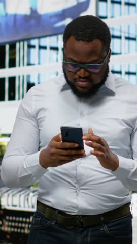 Vertical Video Adult man looks for directions on phone gps and walks across city pavement, modern sense of style and city life. Black person strolling and checking his smartphone apps, urbanism
