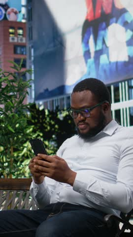 Vertical Video African american man sitting on bench reading online content on his phone, modern lifestyle and expressive attitude highlighted by skyscrapers buildings and the metropolitan energy