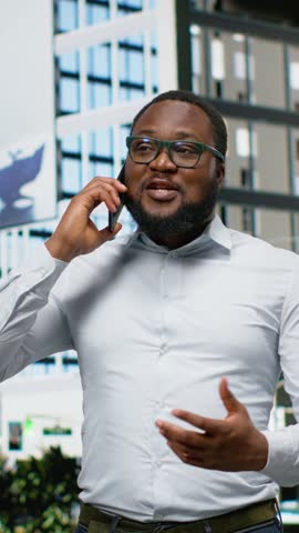 Vertical Video African american adult man on a city street answering a phone call, captures independence and modernity surrounded by sunlight and the scenery of a busy metropolitan environment. Camera