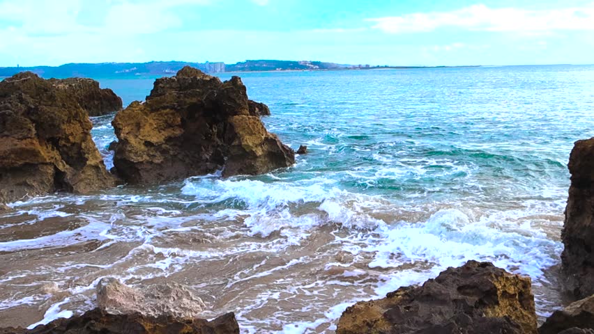 Slow motion handheld footage of foamy water striking sharp rocks and sand along the Caxias shoreline in Portugal with distant hills on the horizon.
