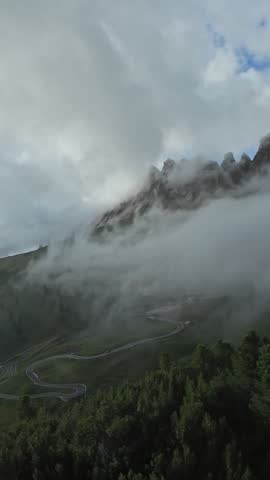 Vertical screen Fog rolling over mountain pass with low clouds hugging jagged peaks and winding road below, mountain guide and weather researcher observing shifting conditions from ridge.