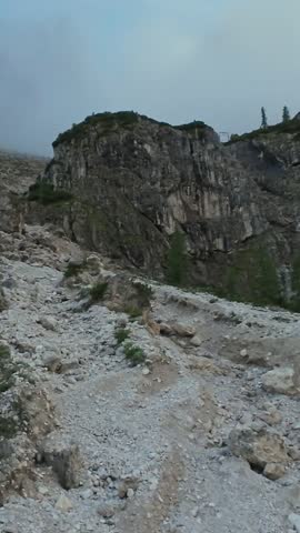 Vertical screen Rugged rocky trail through boulder field under low cloud and mist, loose scree and scattered stones marking route; mountaineer navigating careful steps, cool wind and stark