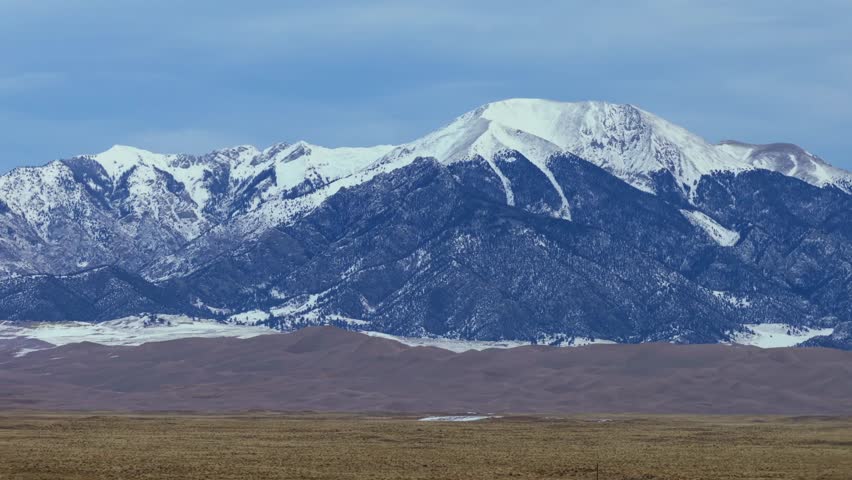 Aerial view of the massive Great Sand Dunes field in the San Luis Valley. The towering snowy peaks of Mount Herard and the Sangre de Cristo Range provide a dramatic backdrop to the desert landscape.