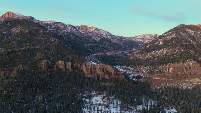 Aerial view of a small convoy of cars driving down the steep winding curves of Wolf Creek Pass. The sunset creates long shadows across the snowy landscape and volcanic rock cliffs.
