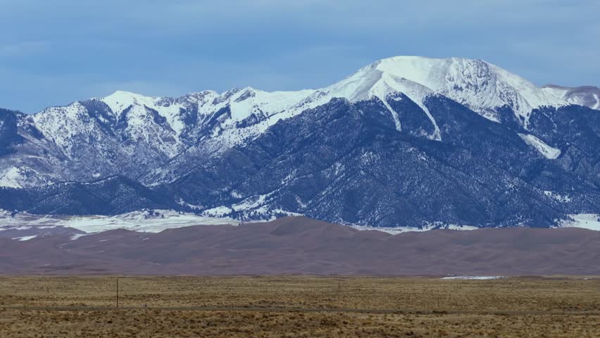 High-angle drone shot capturing the intersection of desert dunes and alpine peaks. The snow-covered slopes of Mount Herard rise sharply above the expansive Great Sand Dunes in Southern Colorado.