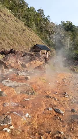 Hot spring water flows, producing thick steam rising into the sky, with a charming gazebo and scenic hills in the background.