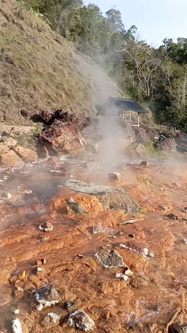 Hot spring water flows, producing thick steam rising into the sky, with a charming gazebo and scenic hills in the background.