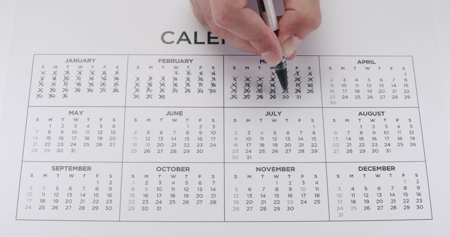 A man's hand marks dates off on a yearly calendar with a black pen. Useful for deadlines, appointments, business, and events. Marking Dates On Calendar
