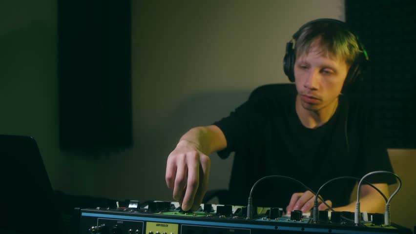 Young male musician with headphones composing a new techno track, meticulously adjusting knobs on an analog synthesizer in a professional sound recording studio with moody green lighting