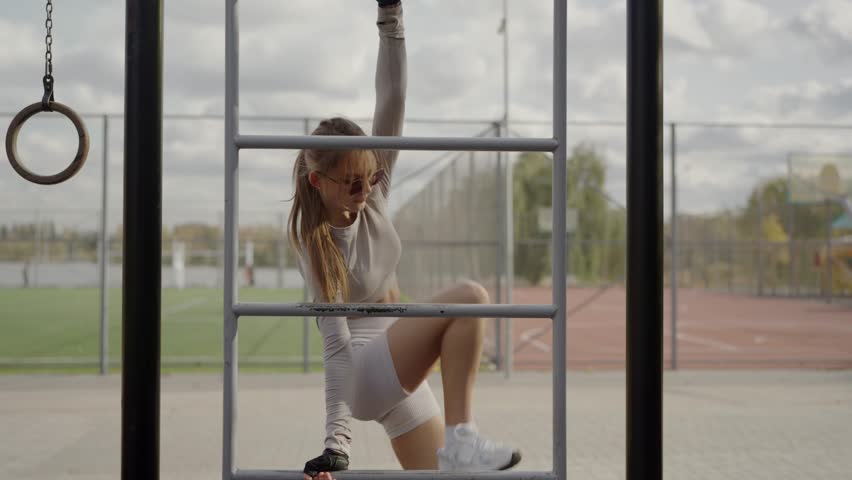 Athletic Woman Performing Aerial Stunts on Outdoor Gym Equipment. Young athletic woman showcases flexibility and strength, hanging upside down on outdoor gym bars on a cloudy day.