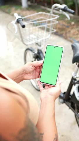 Over the shoulder view of a tattooed woman holding a smartphone with a chroma key green screen, with a bicycle parked outdoors in the background, ready for repair or cleaning instructions