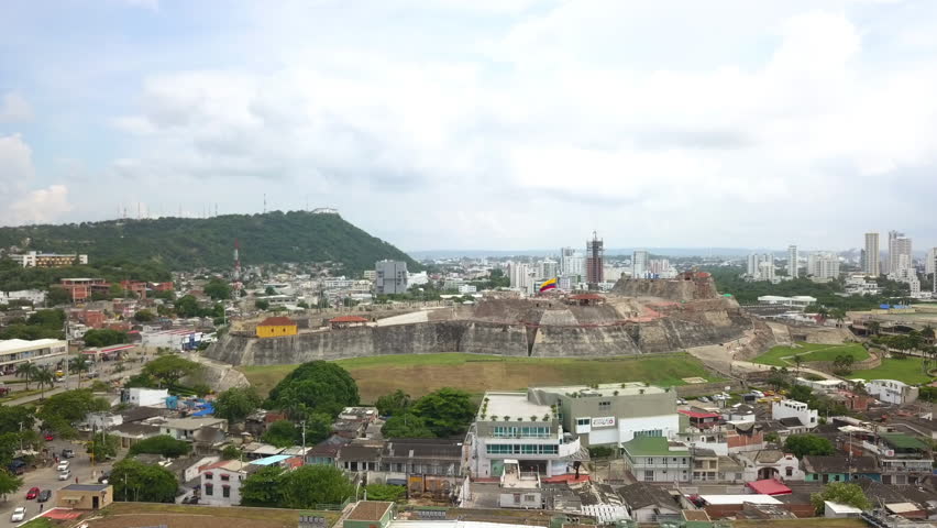 Aerial pan right across historic stone fortress surrounded by urban neighborhoods and modern city skyline under daylight.