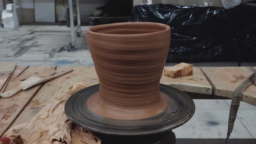 A freshly shaped clay pot sits on a spinning pottery wheel surrounded by sculpting tools in a ceramic workshop. Earthy tones and copy space present