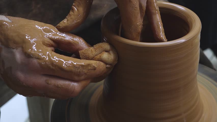 Close-up of a potter smoothing a clay vase using a sponge while shaping it on a spinning wheel. Hands are coated with wet clay, showing detailed craftsmanship